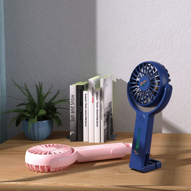 A blue and pink handheld fan with adjustable angles sits on a wooden table beside a potted plant and books, featuring a logo.
