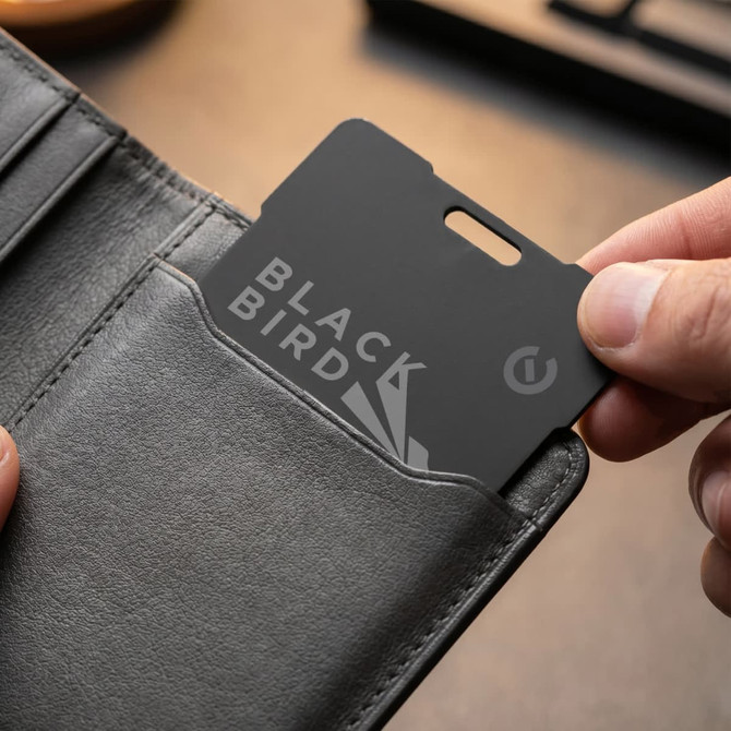 A black Bluetooth tracker being held and placed in a grey wallet pocket. The tracker features a logo.