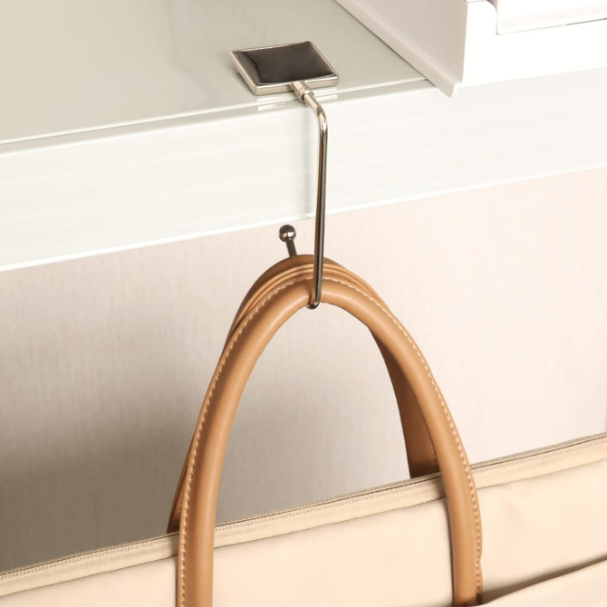 A square silver desk hook holding a beige bag, mounted on a white surface.