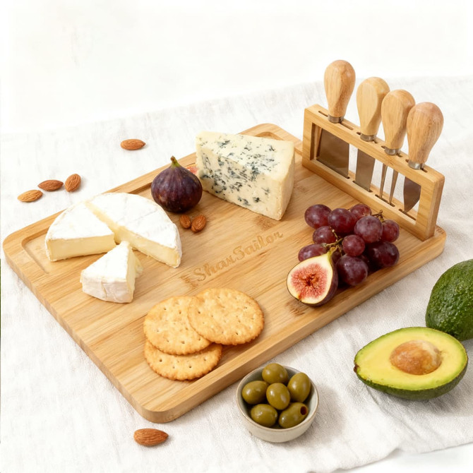 A bamboo cheese board with assorted cheeses, fruits, nuts, crackers, olives, and a knife set with wooden handles.