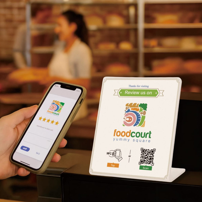 A NFC Review Stand in white, displaying a logo and QR code, beside a person's hand holding a smartphone.