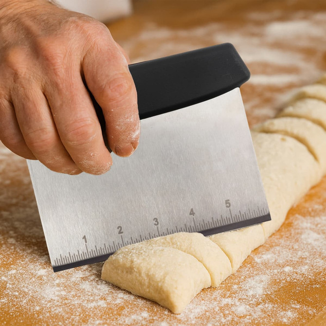 A bench scraper with a black plastic handle, featuring a stainless steel blade, cutting through dough on a floured surface.