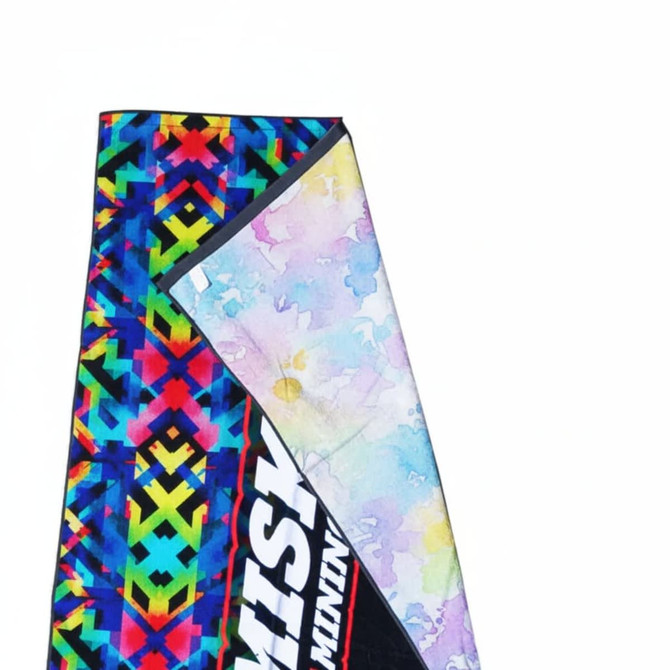 A beach towel featuring a vibrant, multi-coloured geometric pattern with a soft, pastel-lined underside and a logo.
