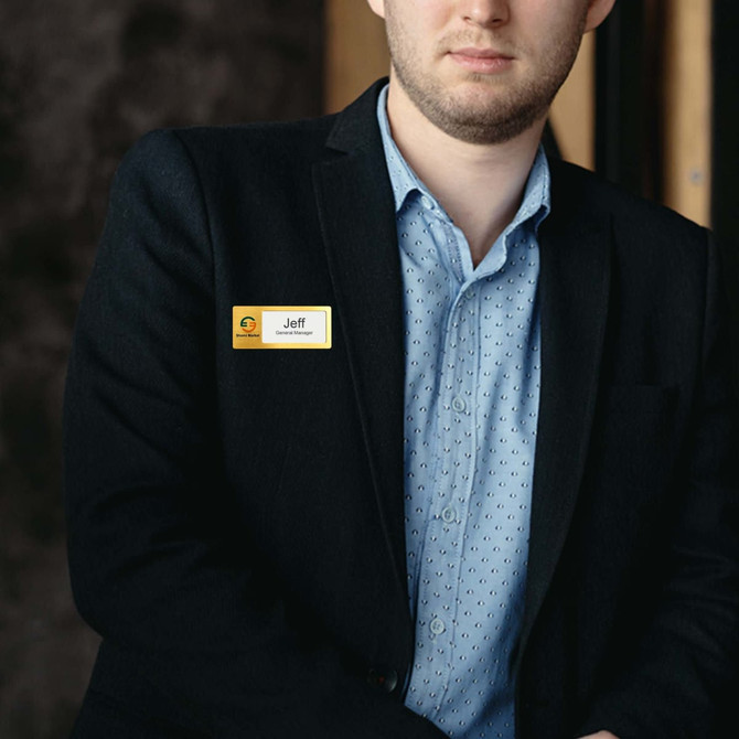A man in a blazer wearing a gold and silver reusable metal name badge. The background is blurred.