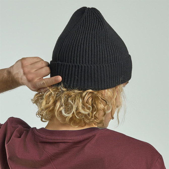 A black knitted beanie displayed on a person with curly hair, viewed from the back. The person wears a maroon shirt.