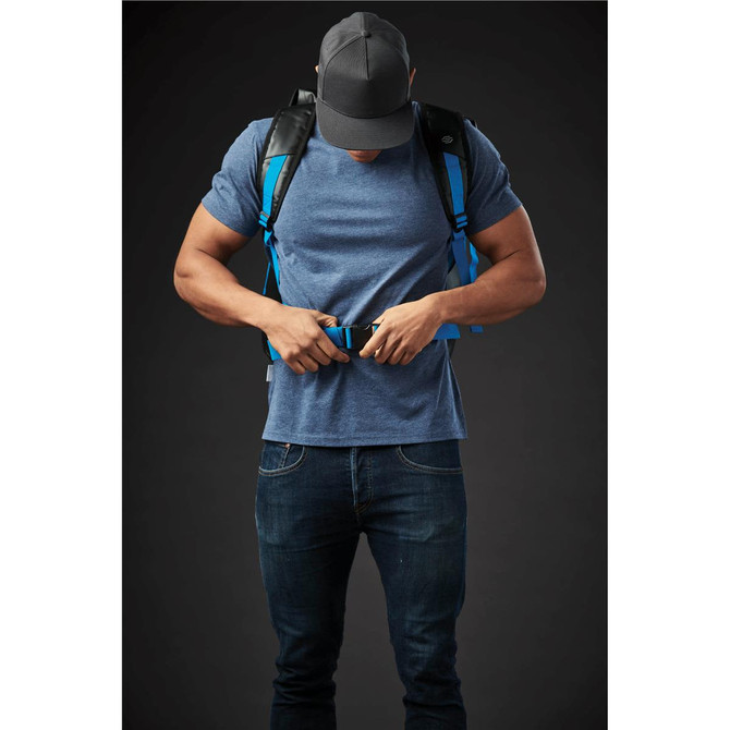 A person adjusts the straps on a blue Kemano Backpack while wearing a grey T-shirt and a cap. The backpack features black accents.