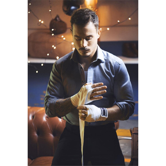 A man in a blue button-up shirt wraps his hands in white tape, surrounded by soft lighting and a casual setting.