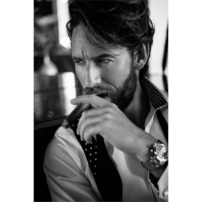 A model with a beard holding a cigar, wearing a white button-up shirt with black accents and a watch.