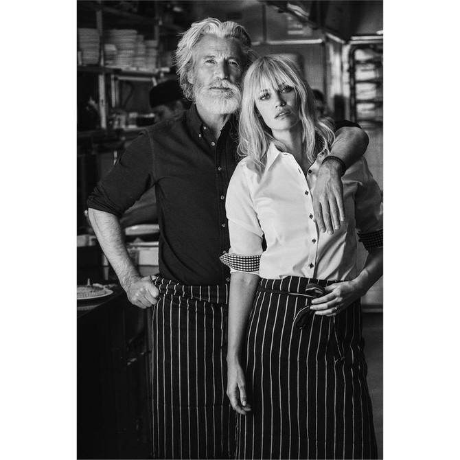A man and a woman pose together in a kitchen setting, both wearing stylish button-up shirts. The woman’s shirt is white, and the man's is dark.