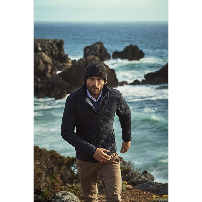 A man wears a navy quilted jacket and a beanie, walking along a rocky shoreline with ocean waves in the background.