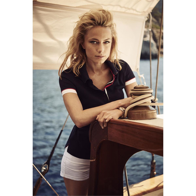 A women's cotton polo in navy with contrasting collar, featuring a logo, set against a nautical background.
