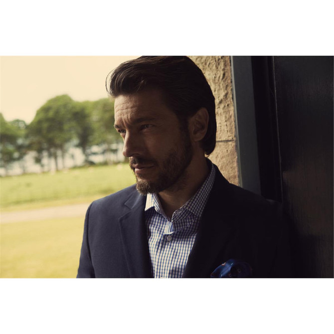 A man wearing a navy button-up shirt with a checkered pattern, standing in front of a door with a landscape background.