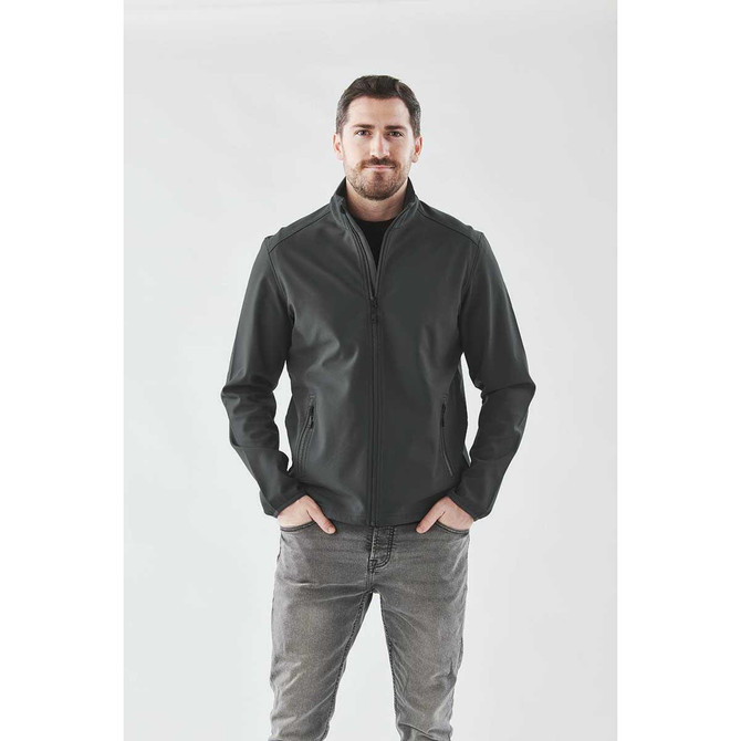 A man models a dark green softshell jacket with a stand-up collar, featuring a logo on the chest.