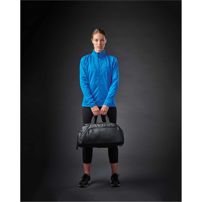 A woman wearing a bright blue lightweight softshell jacket holds a black duffel bag against a dark background.