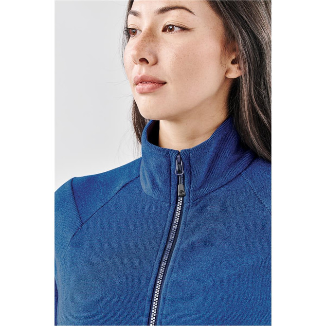 A close-up of a woman wearing a blue full zip jacket, featuring a high collar and a zip closure. It has branding.