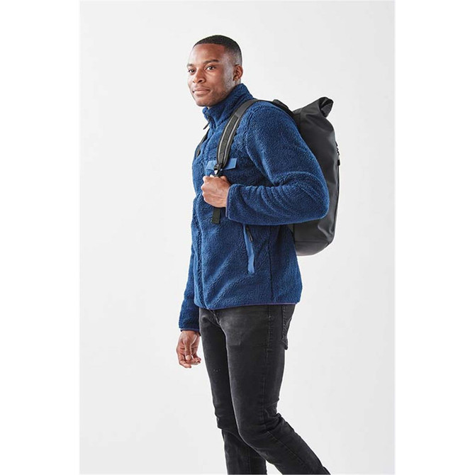 A man in a navy fleece jacket carries a black Sargasso Backpack over his shoulder against a plain background.