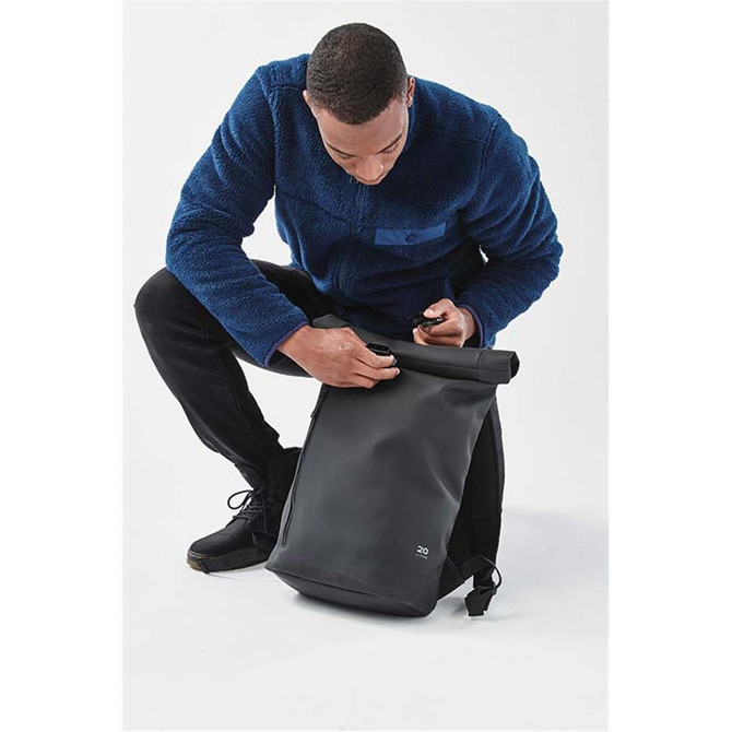 A person in a blue fleece jacket is using a black Sargasso Backpack. The backpack features a roll-top closure and has a logo.