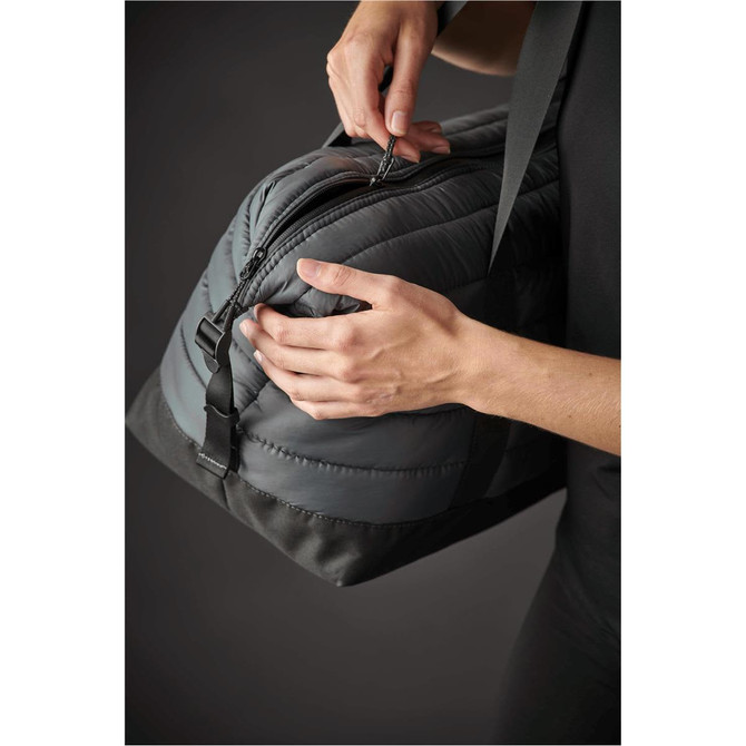 A quilted duffle bag in grey with a zippered opening, being held by hands. Features a logo on the side.