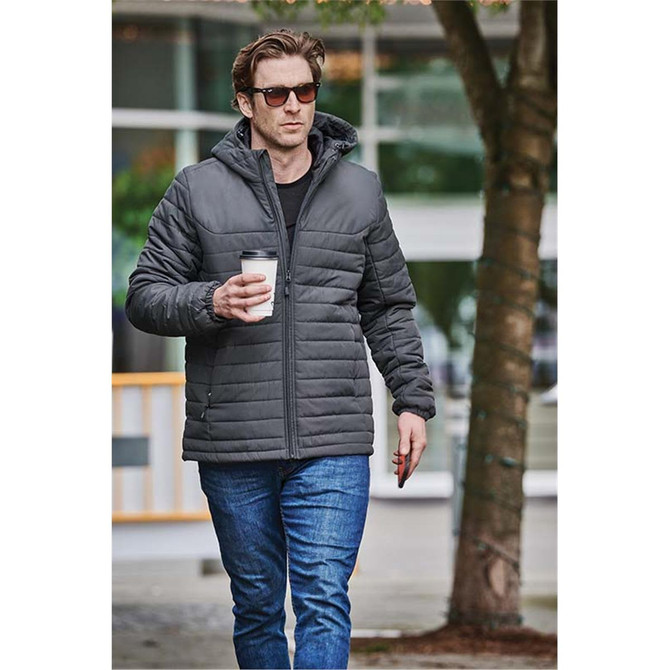 A man in a grey quilted hoody holds a coffee cup, walking casually outdoors in jeans and sunglasses.