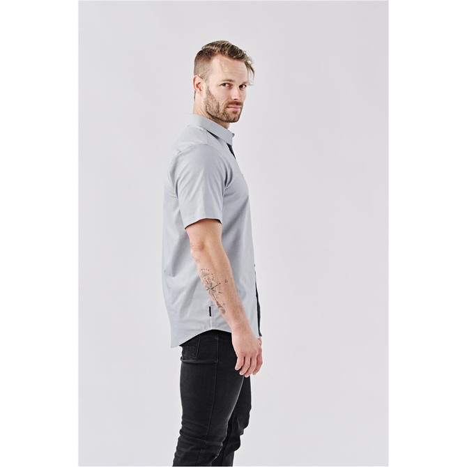 A man wearing a short-sleeved, light grey button-up shirt, featuring a logo, posed against a plain background.