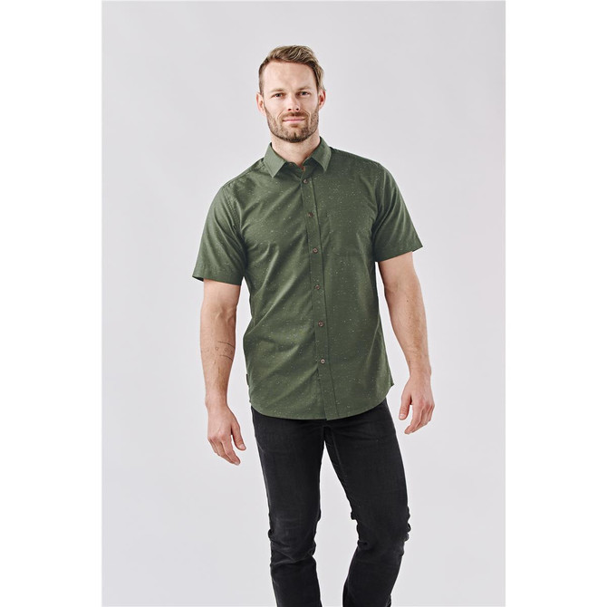 A button-up shirt in dark green with a subtle pattern, featuring a logo, worn by a model with light hair.