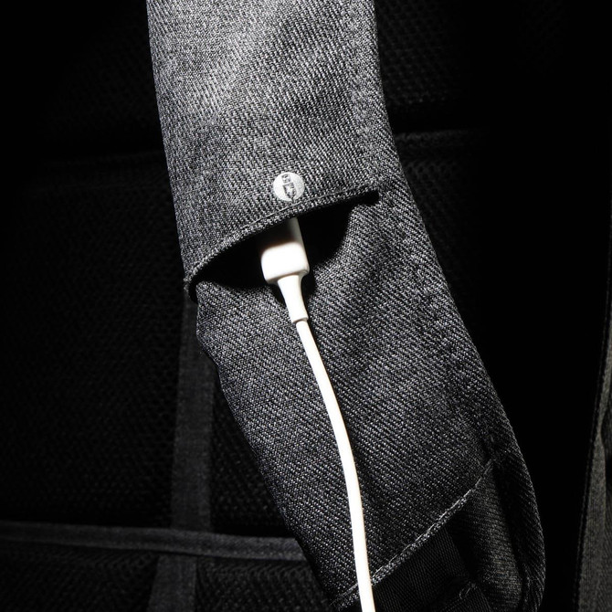 A close-up of a black backpack strap featuring a white charging cable tucked inside. The strap has a logo.