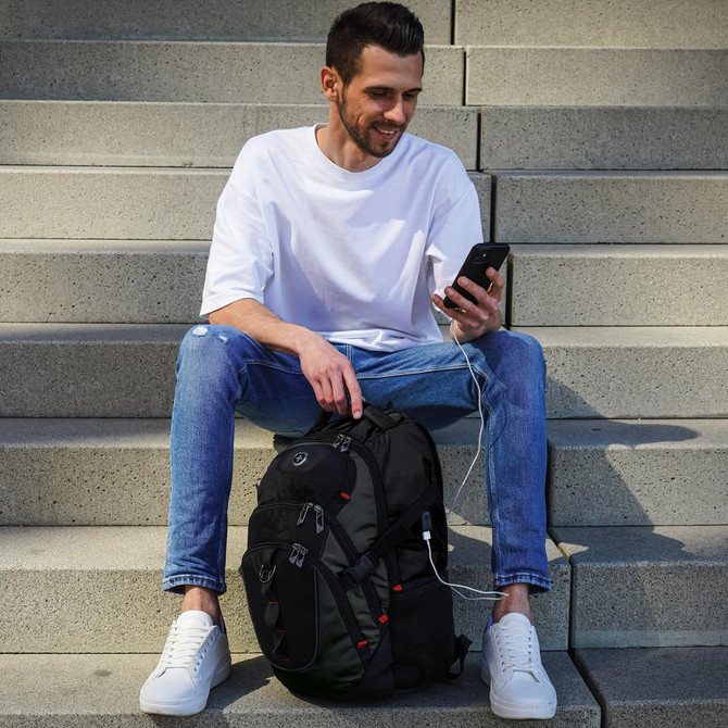 A man sits on steps, holding a phone, with a black Swissdigital Vector Backpack beside him.