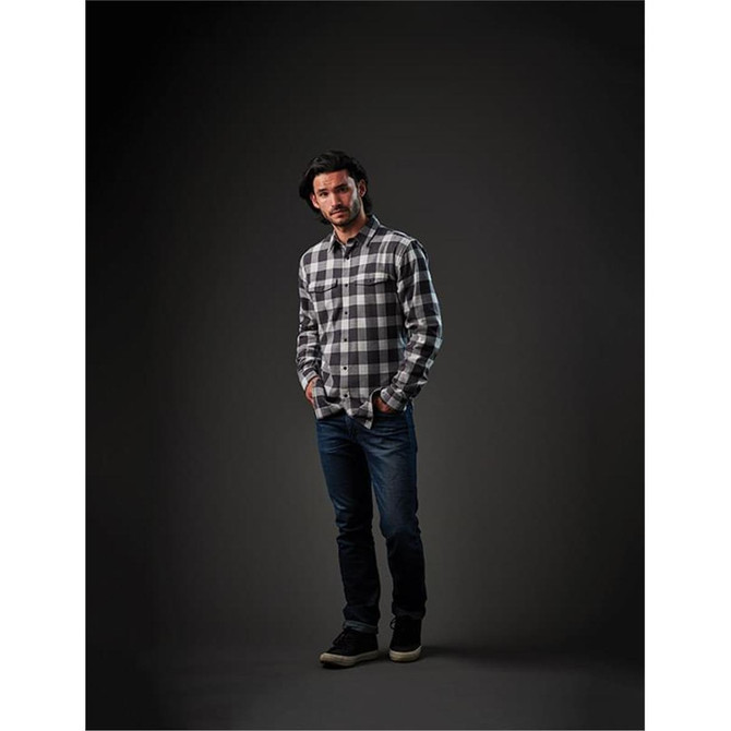 A man stands in a dark setting wearing a black and white checked snap front shirt and blue jeans.