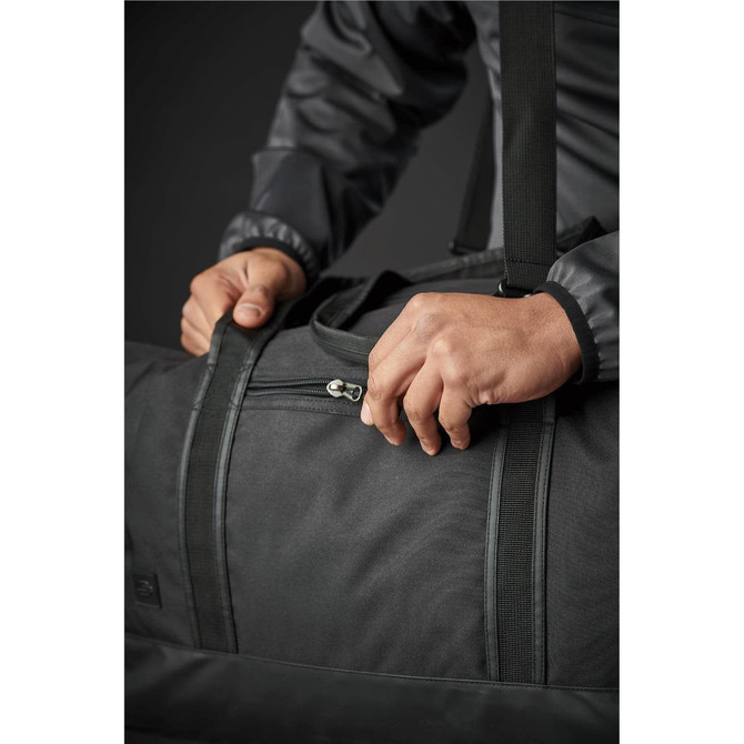 A dark green duffle bag with a zippered opening, being held by a person with a grey jacket. Features a logo.