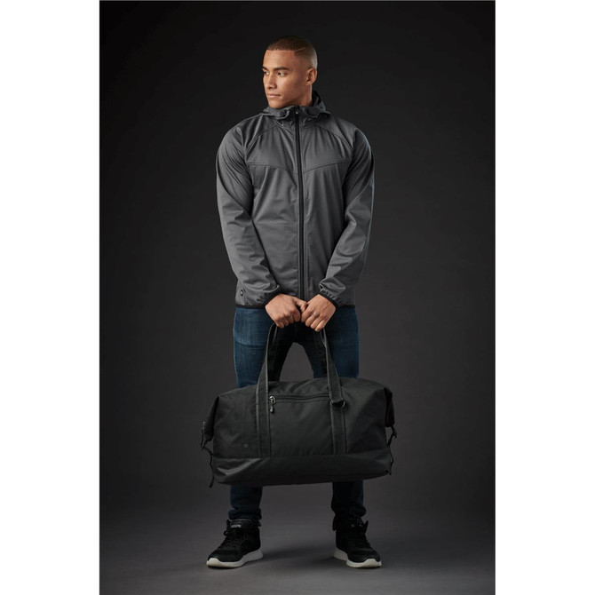 A man wearing a grey jacket holds a black duffle bag with handles, set against a dark background.