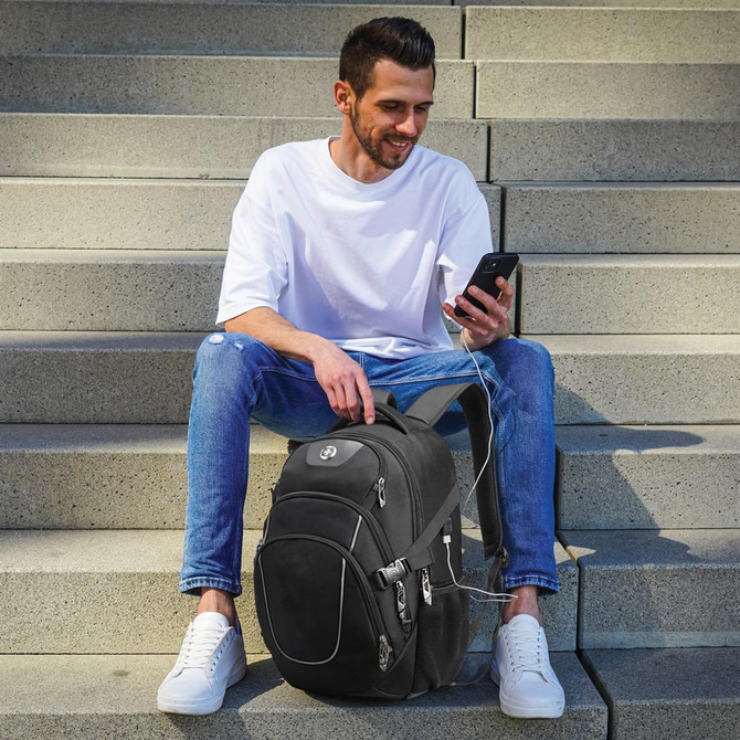 A backpack in black, equipped with a charging port, rests beside a man seated on steps, using a smartphone.