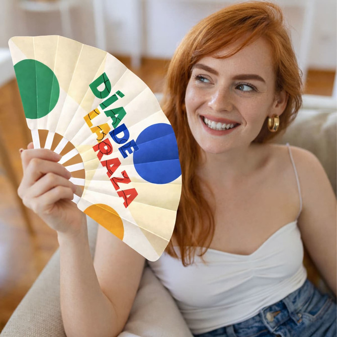 A hand fan featuring colourful circles in green, orange, and blue, held by a smiling person with red hair.