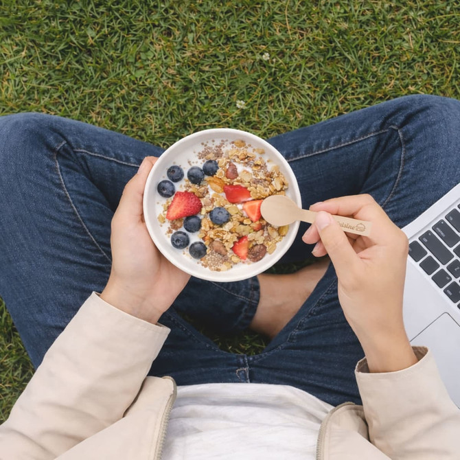 A person holding a bowl of granola topped with berries, using a small wooden disposable spoon, sitting on grass.