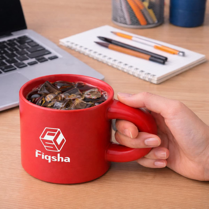 A red ceramic coffee mug being held, filled with iced drink, alongside a laptop and stationery items on a desk.