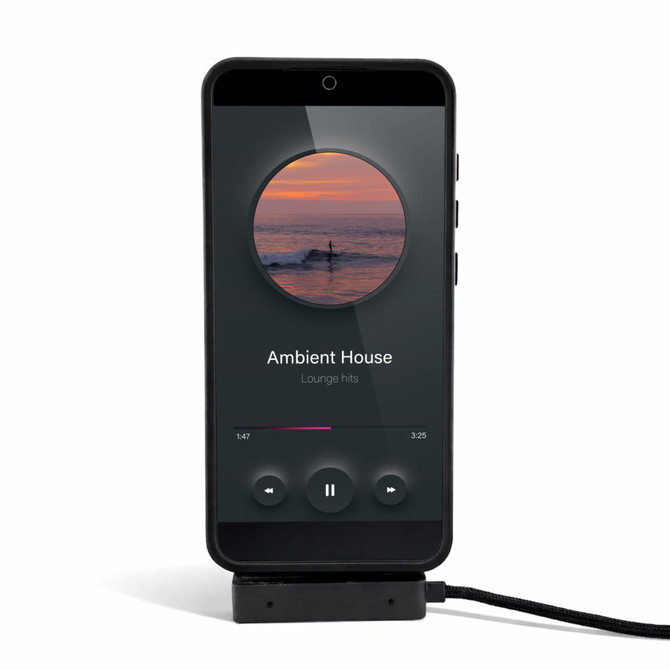 A black phone on a stand displaying a music player interface with a charging cable attached.