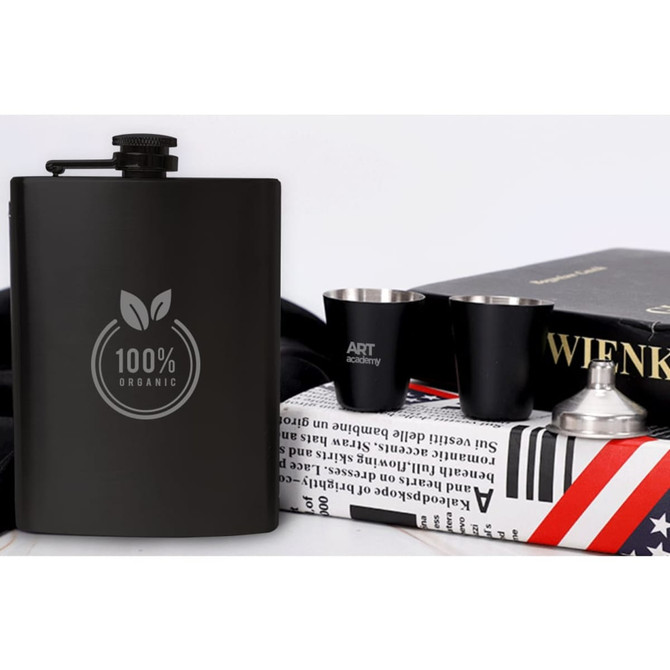 A black stainless steel hip flask set featuring two shot cups and a funnel beside books and a black cloth.