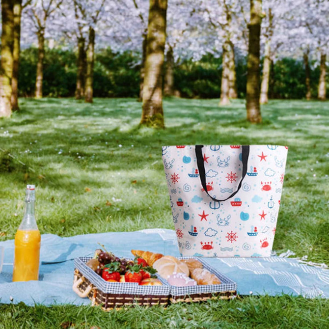 A large cooler tote bag in white with colourful nautical patterns, alongside a picnic spread and a drink bottle.