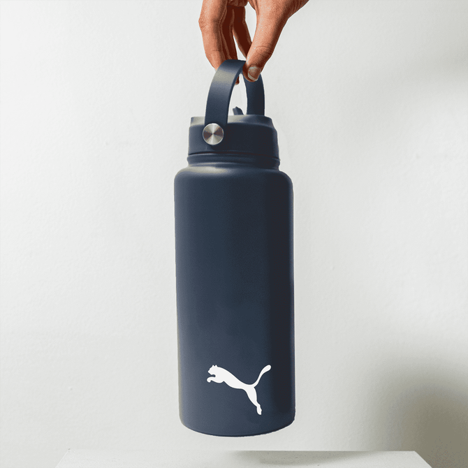 A drink bottle in dark blue with a handle and a logo on the front, suspended by a hand.
