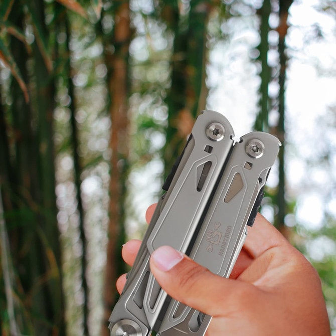 A silver survival multi-tool is held in a hand, set against a background of blurred bamboo. It has a logo on it.