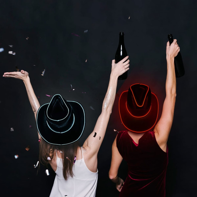 Two women wearing black cowboy hats with glowing blue and red LED lights, celebrating with drinks in hand amid confetti.