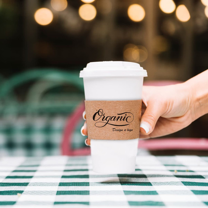 A white compostable paper cup with a brown cup sleeve held by a hand, set on a checkered table.