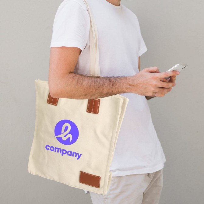 A cream-coloured canvas bag with brown accents and a logo, held by a person checking their phone.