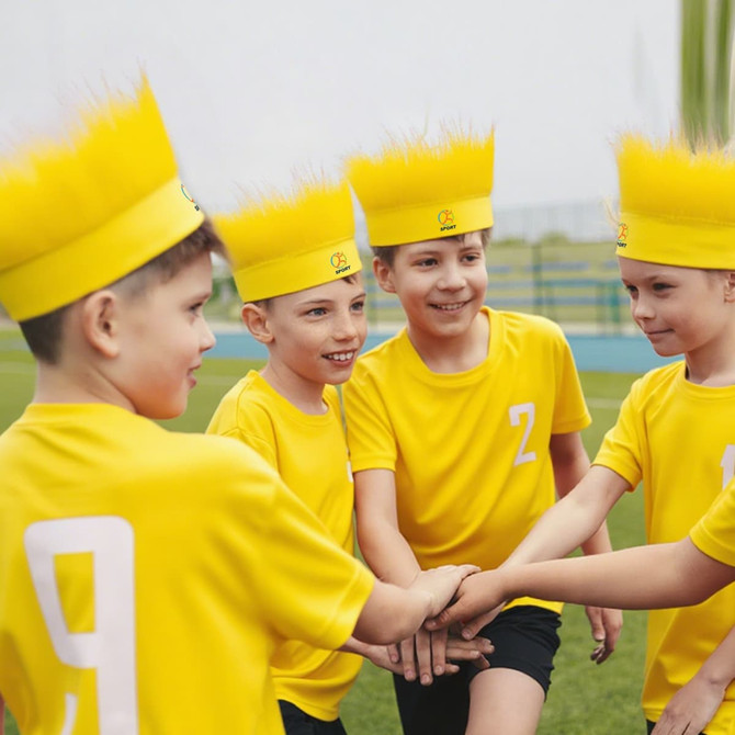 Five boys wearing bright yellow shirts and fluffy yellow crowns are engaging in a playful group activity outdoors.