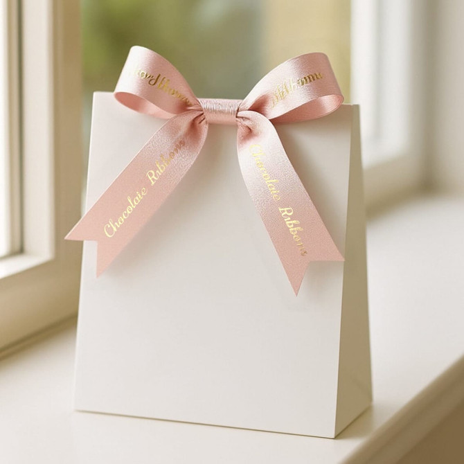 A white gift bag adorned with a soft pink satin ribbon bow, featuring shiny gold lettering.