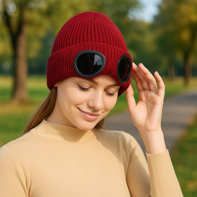 A red beanie with black goggles worn by a smiling woman in an outdoor setting.