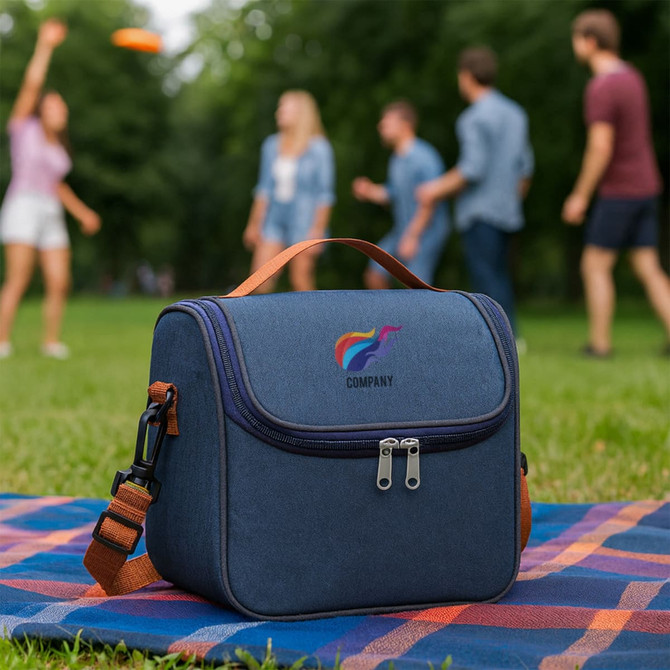 A blue customizable lunch bag with a strap and a logo, sitting on a picnic blanket in a park.