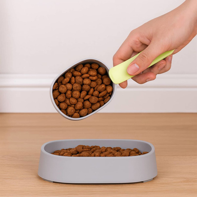 A grey pet food bowl filled with kibble, being scooped with a green and grey 2 in 1 pet food scoop.