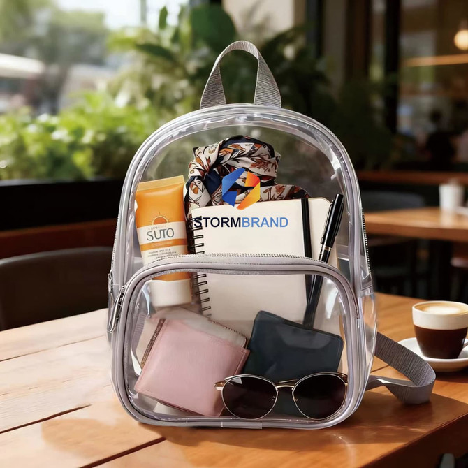 A clear mini backpack with various items inside, including a notebook, wallet, sunglasses, and a skincare product, with a logo.