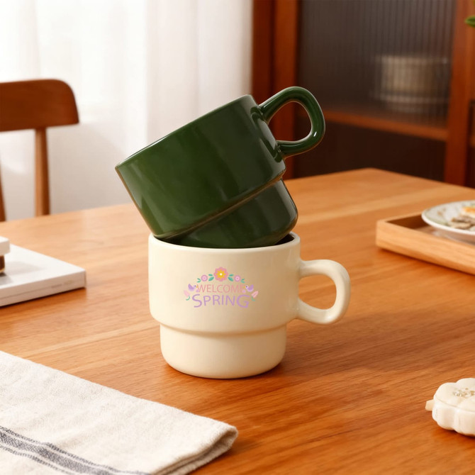 A stackable ceramic mug set in green and cream sits on a wooden table, featuring a branding logo.