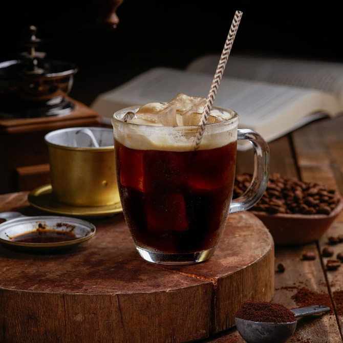 A glass of dark iced coffee with a cream top, featuring a brown-striped paper straw, sits on a wooden table.